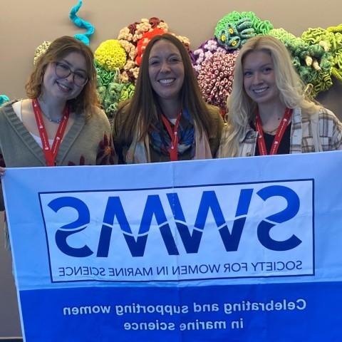 Three UNE researchers pose holding a Society for Women in Marine Science banner